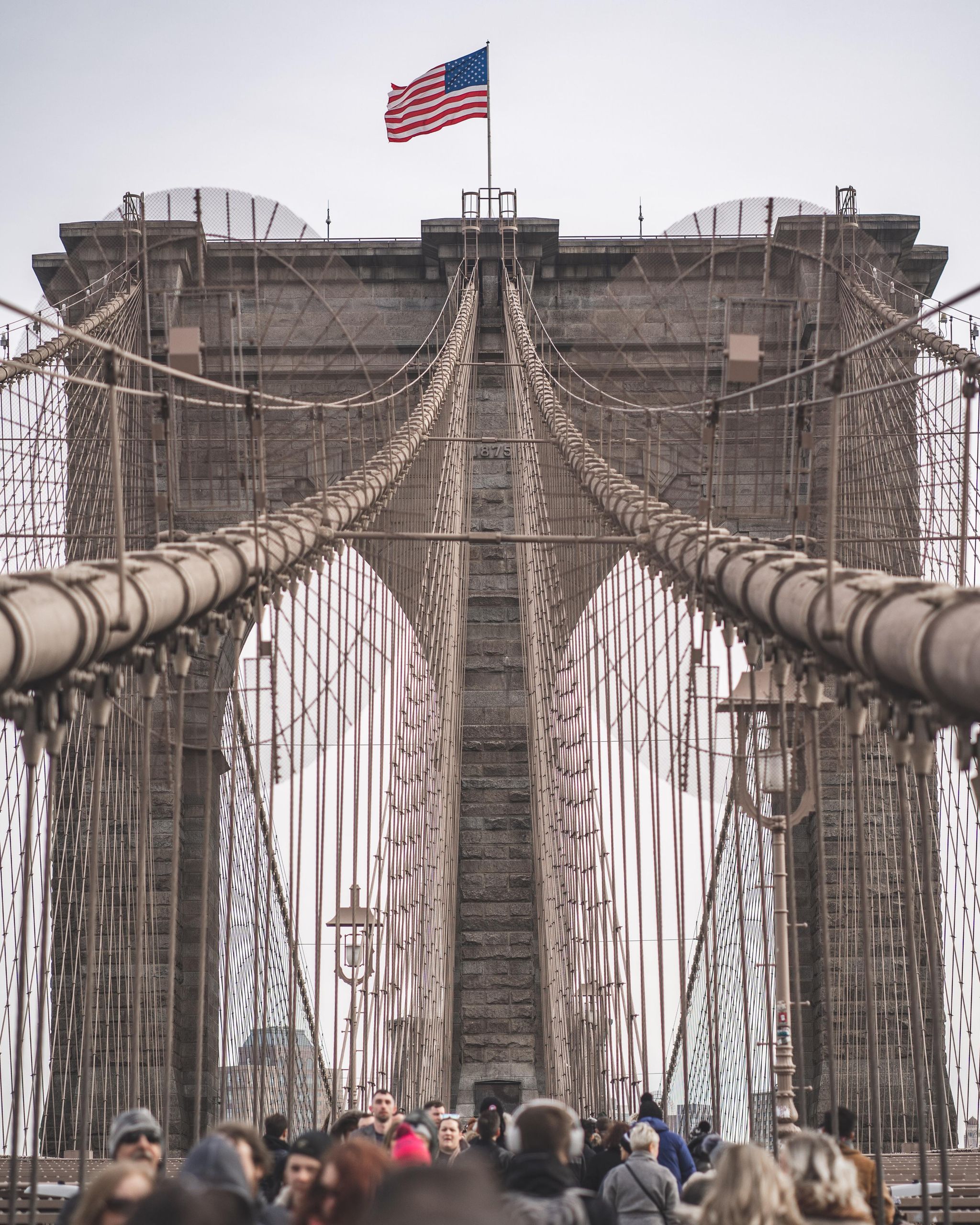 View of the Brooklyn Bridge with people walking and an American flag waving, capturing NYC's essence.