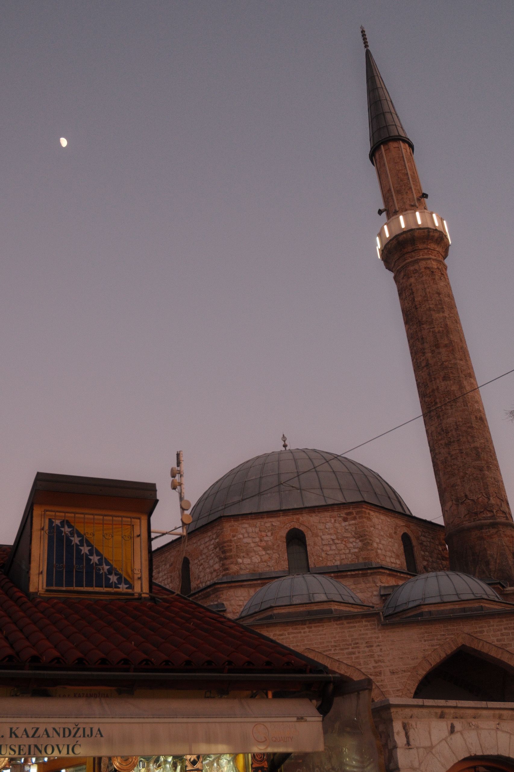 View of the Gazi Husrev-beg Mosque minaret and dome in Sarajevo at twilight with the moon visible.